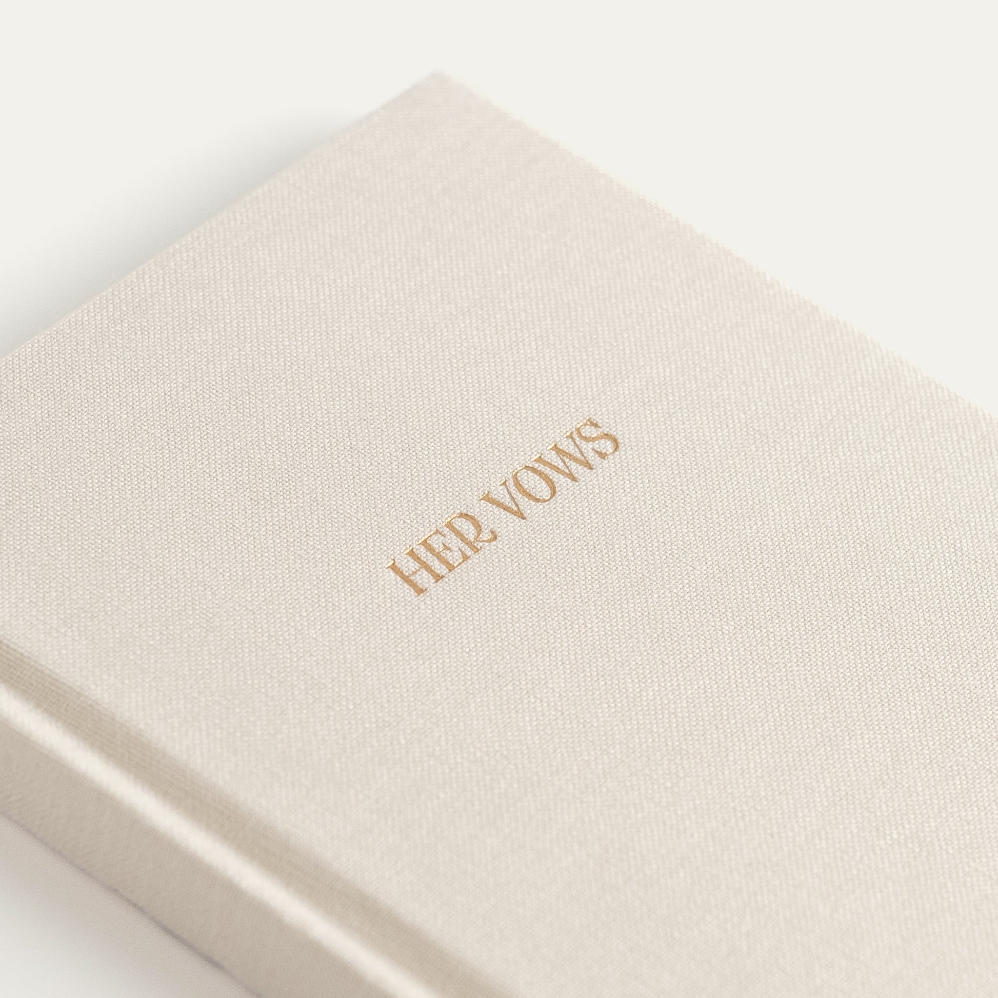 A close-up of a cream-colored textured hardcover book or journal with the title 'HER VOWS' printed in gold foil on the cover, set against a clean white background.