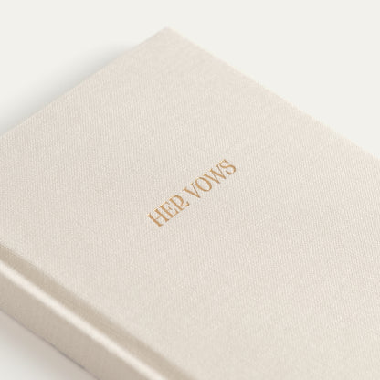 A close-up of a cream-colored textured hardcover book or journal with the title 'HER VOWS' printed in gold foil on the cover, set against a clean white background.